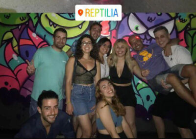 Grupo de 9 amigos sonríen de noche a cámara contra un fondo lleno de dibujos de monstruos y lagartos graciosos